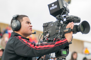 Student wearing a headset and filming an outdoor sporting event with a professional camcorder.
